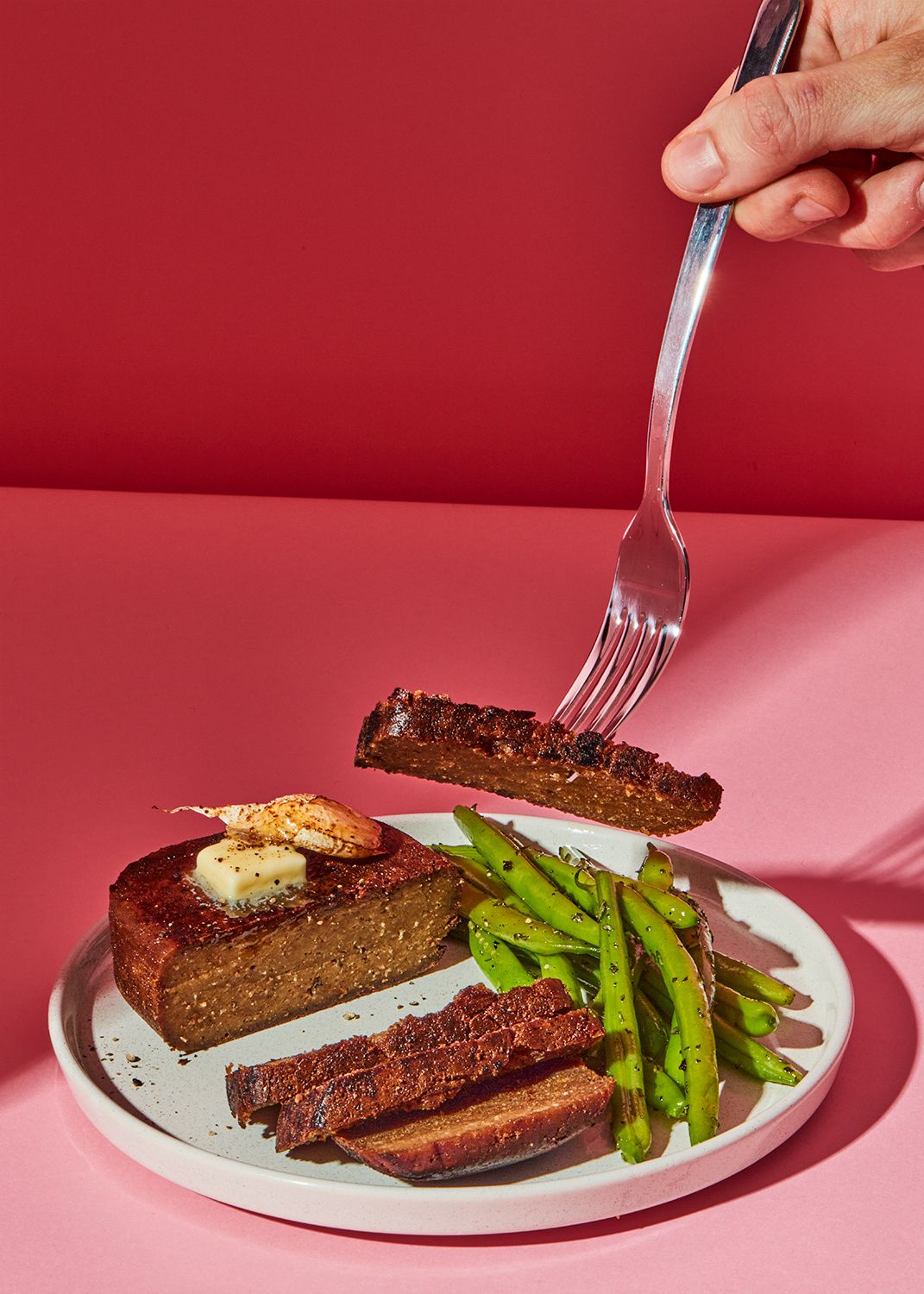 Zubereitetes Seitan-Steak mit Kräuterbutter und grünen Bohnen auf einem Teller vor rosa Hintergrund