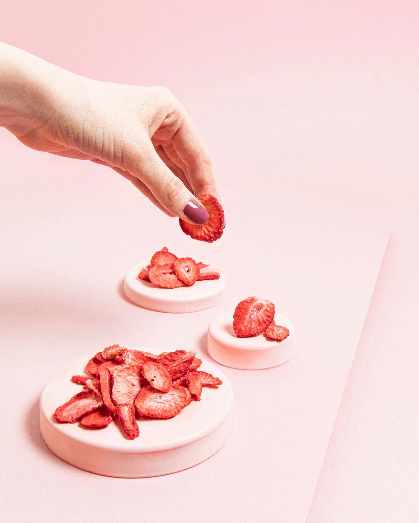 Eine Hand mit lila lackierten Fingernägeln hält eine gefriergetrocknete Erdbeerscheibe über drei kleine, rosa Podeste. Auf den Podesten sind weitere getrocknete Erdbeerstücke vor einem durchgehend rosa Hintergrund angerichtet.