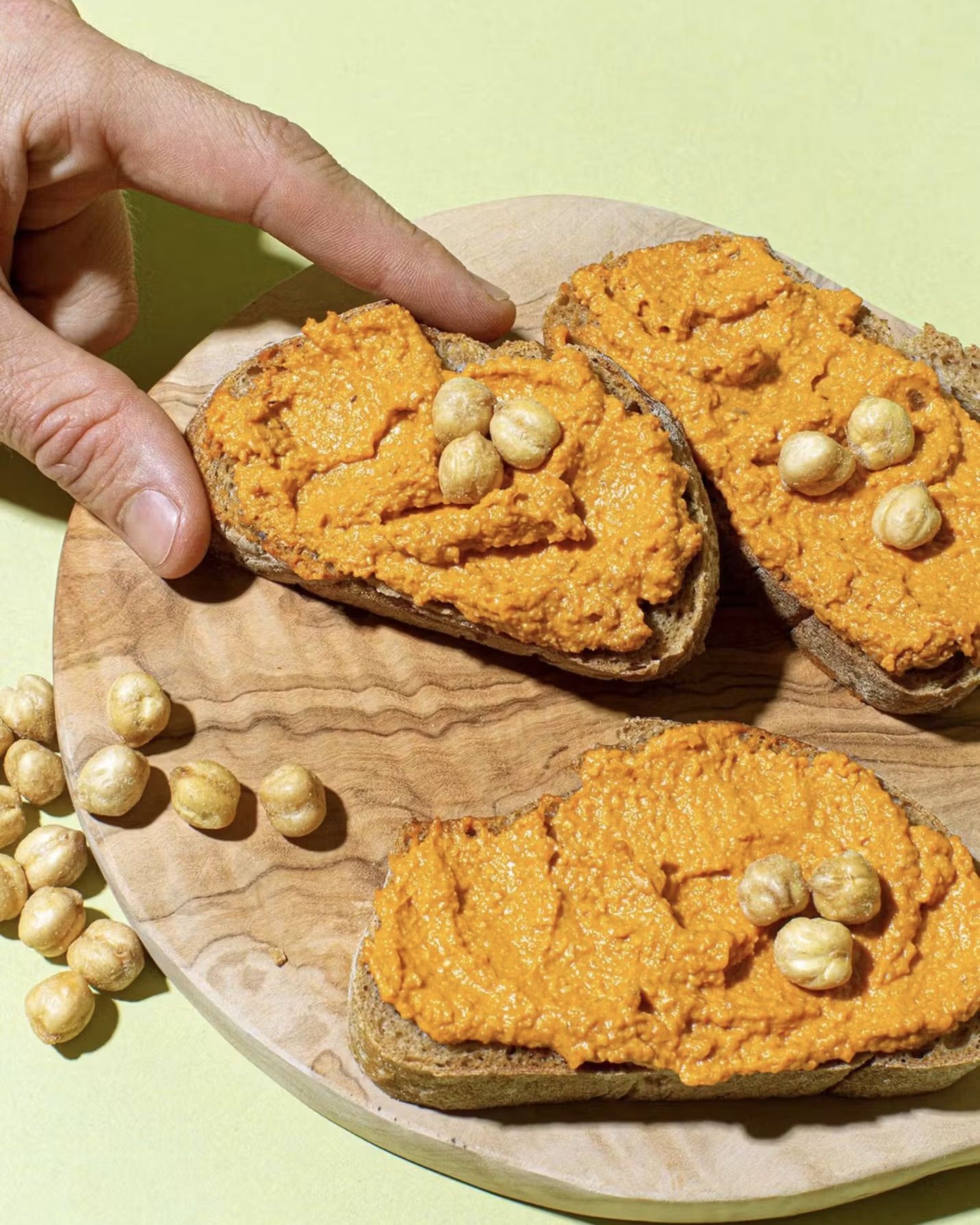 Eine Hand greift nach einer Scheibe Brot mit cremiger Paprika-Kichererbsen-Creme und Kichererbsen-Topping auf einem runden Holzbrett.