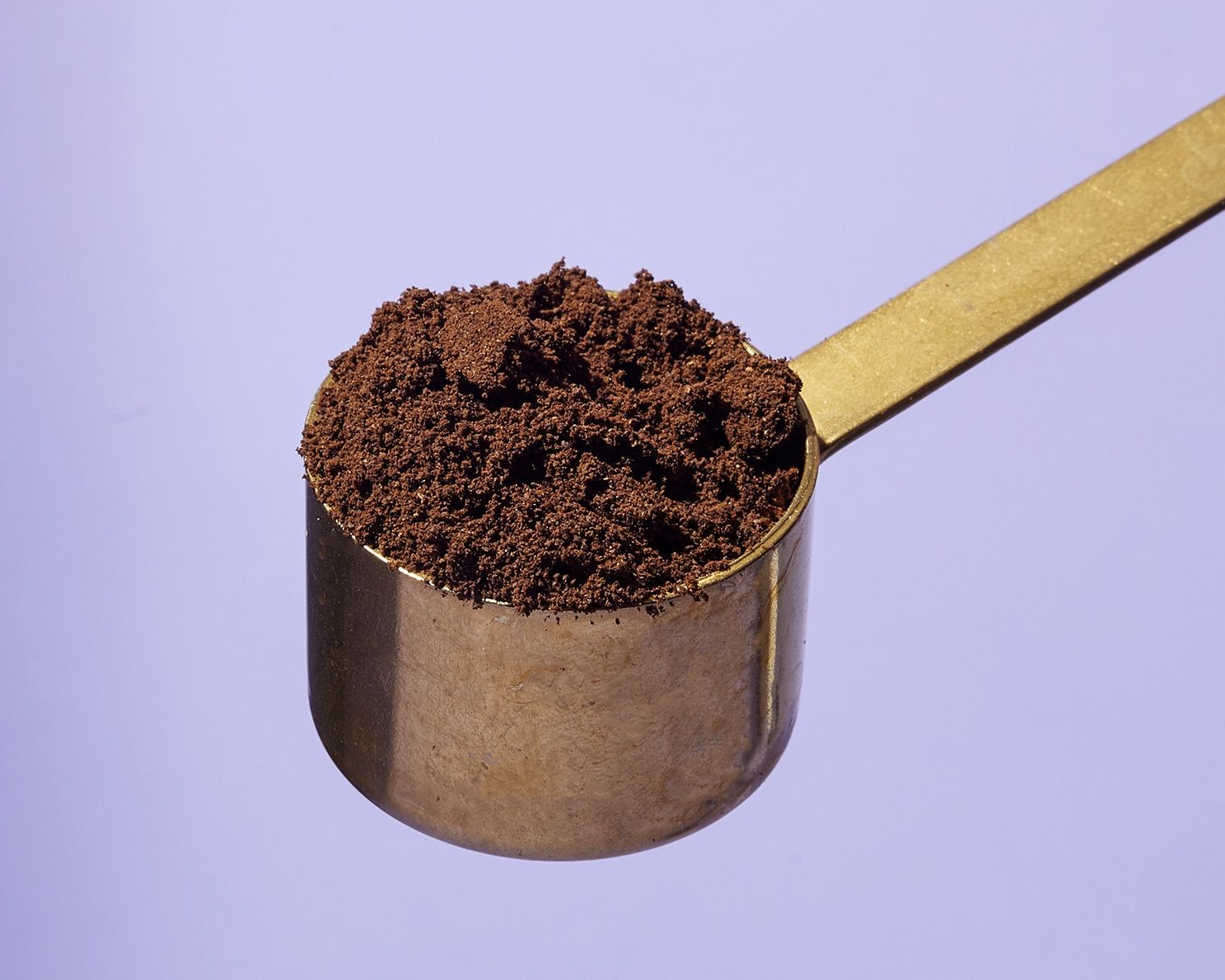 Ein Messlöffel aus Messing voll mit gemahlenem Kaffee für die Zubereitung, Nahaufnahme vor lila Hintergrund.