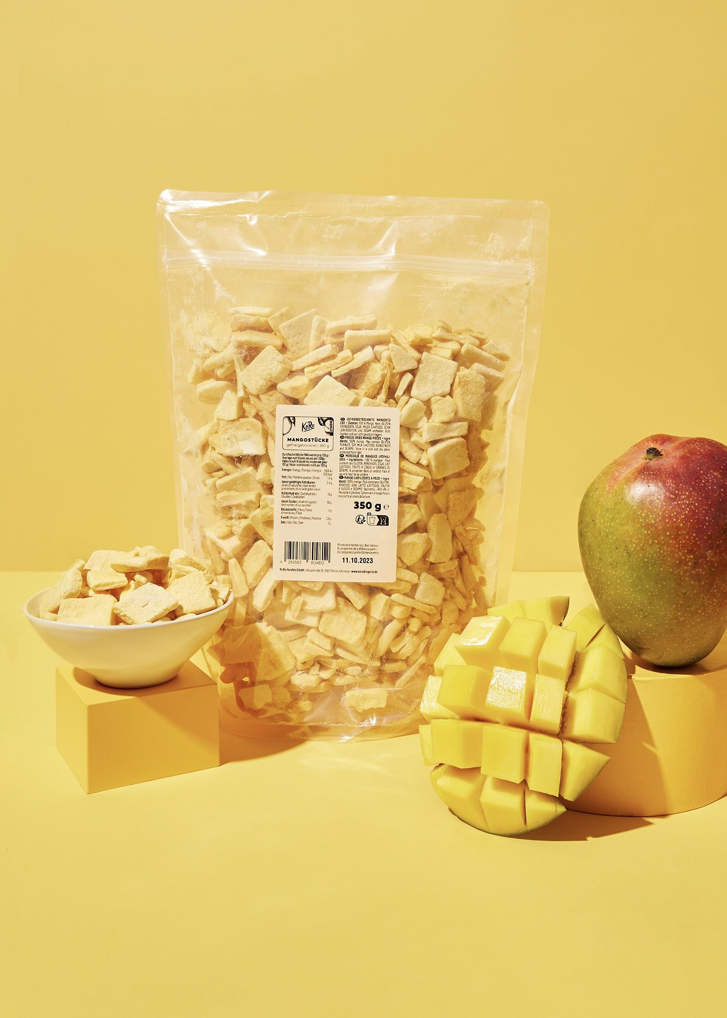 Transparenter Beutel mit gefriergetrockneten Mangostücken von KoRo vor gelbem Hintergrund mit frischer Mango und gefüllter Snack-Schale