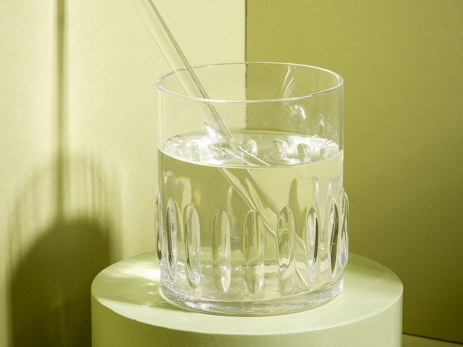Vor hellgrünem Hintergrund steht ein Glas mit einer durchsichtigen Flüssigkeit – darin ein Glasstrohhalm.