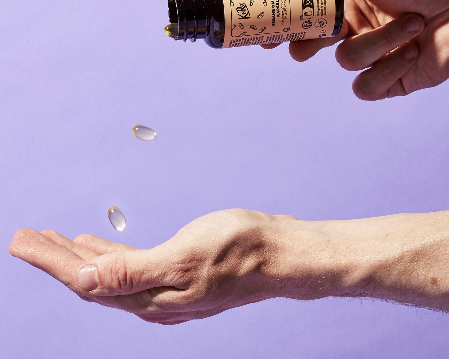 Vegane Omega-3 Kapseln von KoRo fallen aus einer Braunglasflasche in eine geöffnete Handfläche vor lila Hintergrund.
