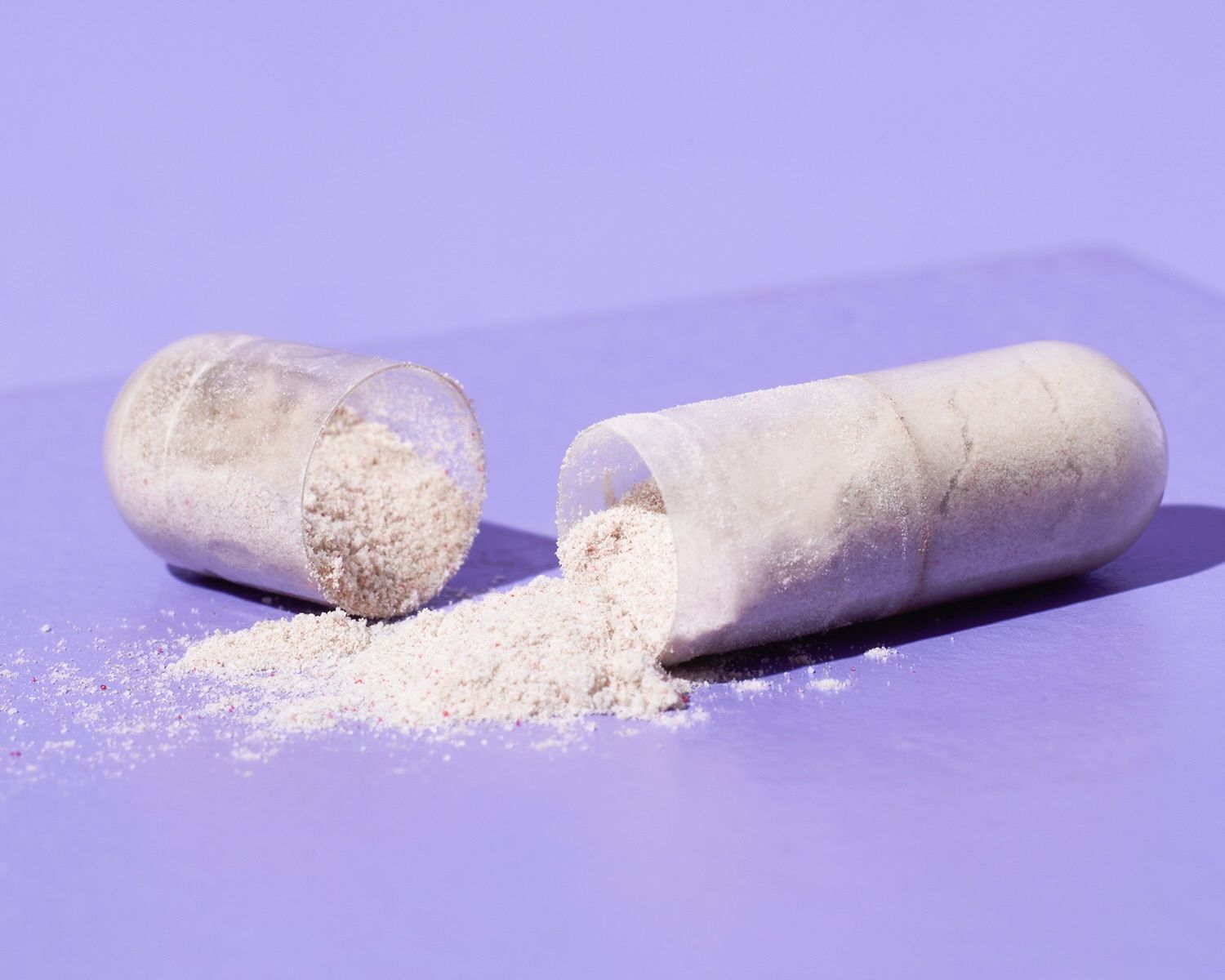 Geöffnete transparente Kapsel mit Eisenbisglycinat und Vitamin C aus Acerola, aus der feines Pulver auf einen violetten Untergrund rieselt.