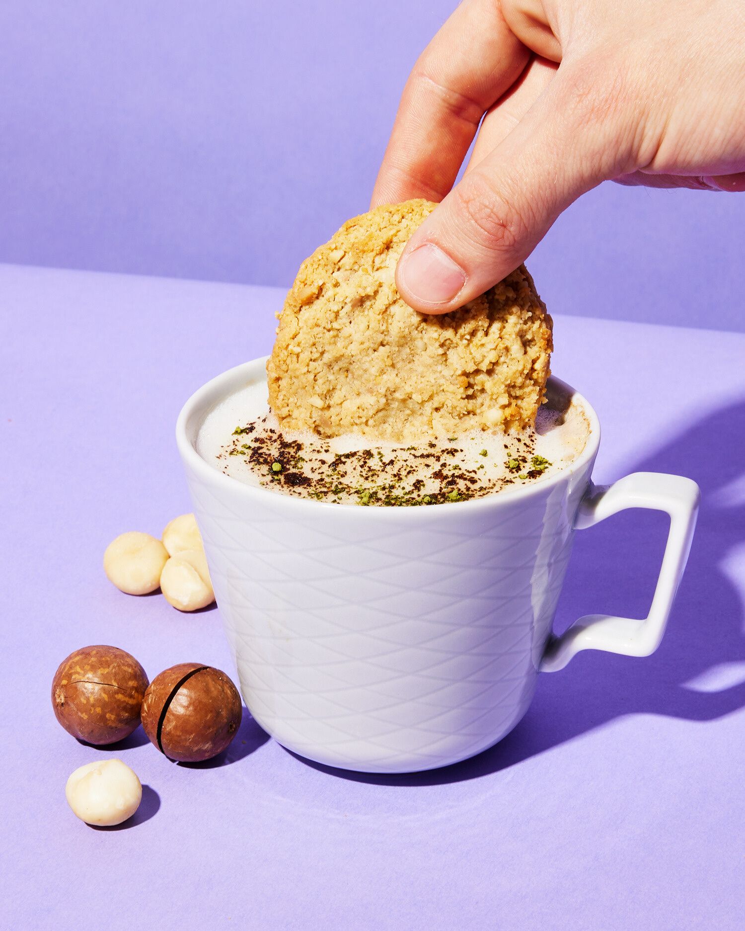 Eine Hand taucht einen runden, goldbraunen Keks in eine weiße Tasse, die ein schaumiges, mit Puder bestreutes Getränk enthält. Neben der Tasse auf einem lila Hintergrund liegen mehrere Macadamianüsse: einige geschält und weiß, andere noch in ihrer braunen Schale.