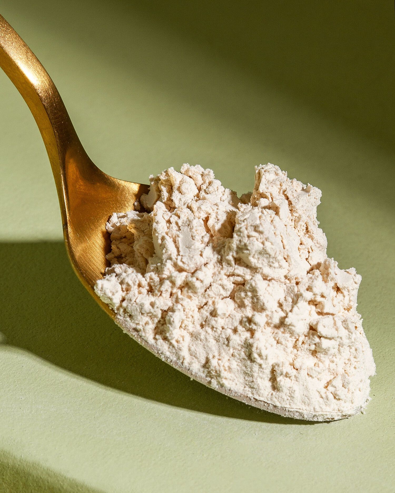 Nahaufnahme eines vollen goldenen Löffels mit hellem, lockerem Proteinpulver, der vor einem hellgrünen Hintergrund liegt. 