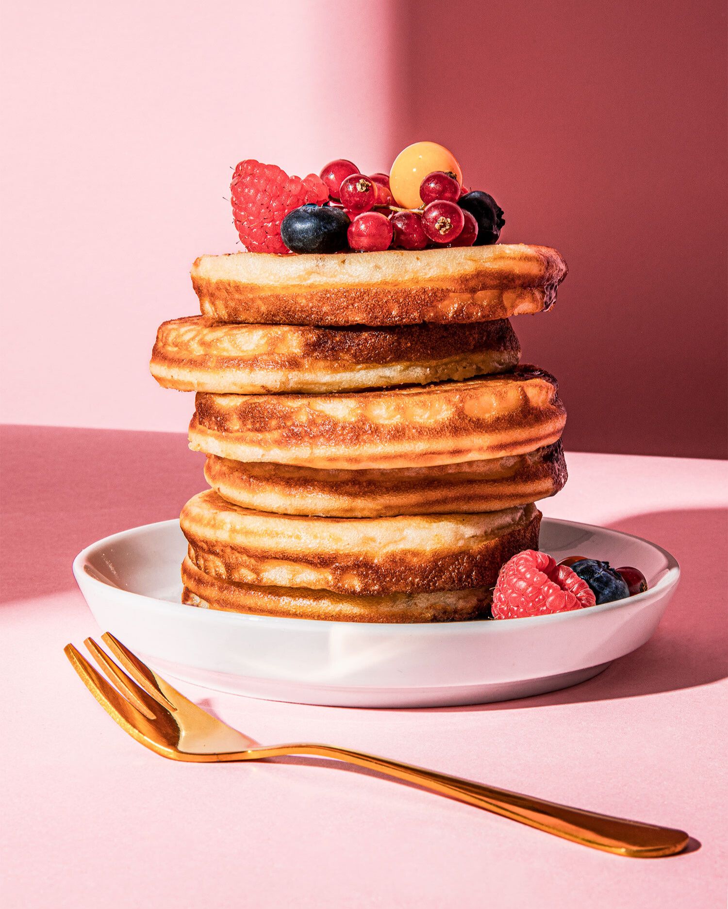 Ein hoher Stapel von goldbraun gebackenen Protein-Pancakes auf einem weißen Teller. Die oberste Schicht ist reichlich mit frischen Beeren dekoriert, darunter Himbeeren, Blaubeeren und rote Johannisbeeren. Vor dem Teller liegt eine goldene Gabel. Der Hintergrund ist in einem leuchtenden Rosa gehalten, mit starken Schattenwürfen.