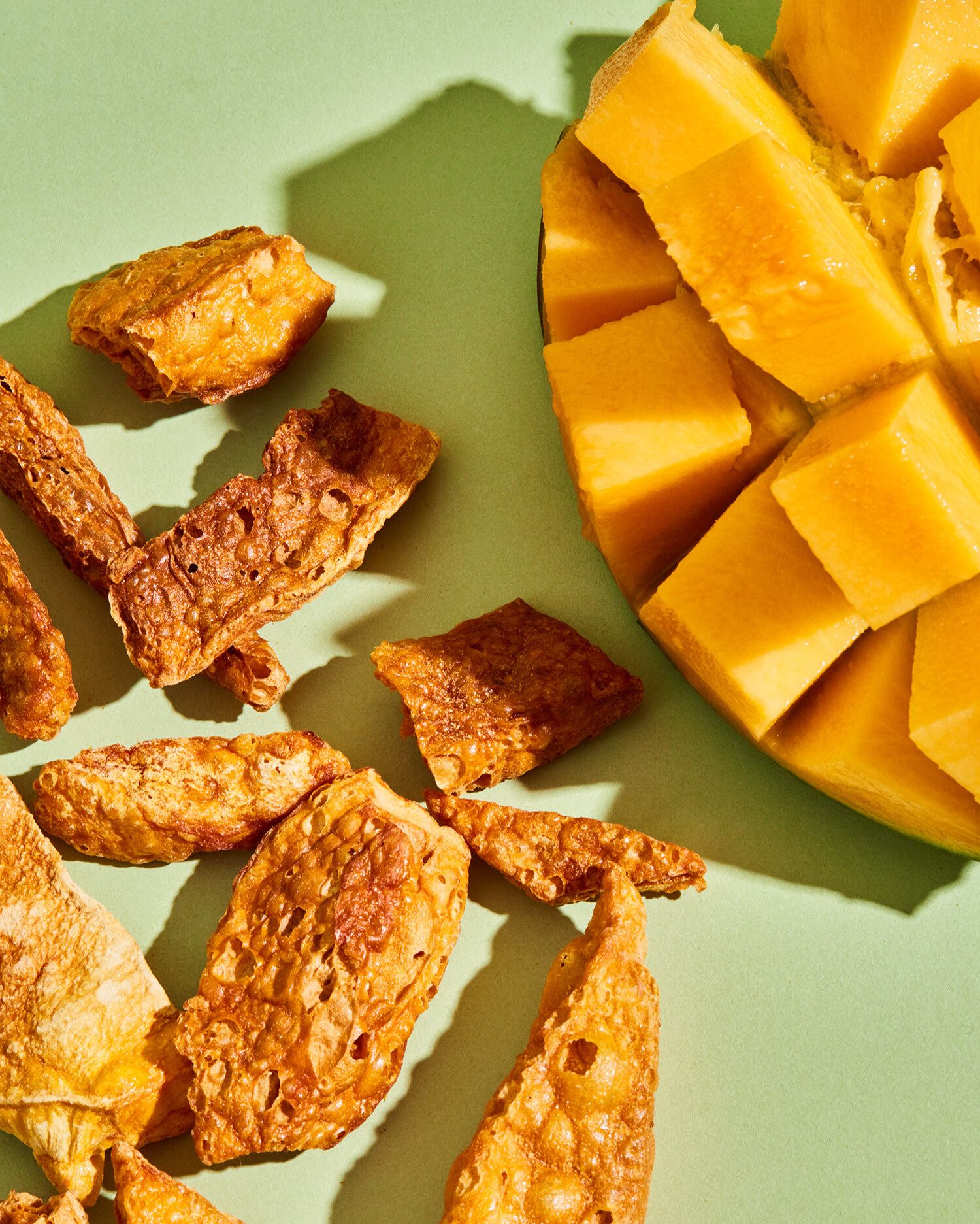 Aufsicht einer halben, in Würfel geschnittenen, frischen Mango neben mehreren goldbraunen, knusprig gepufften Mangostücken auf hellgrünem Untergrund.