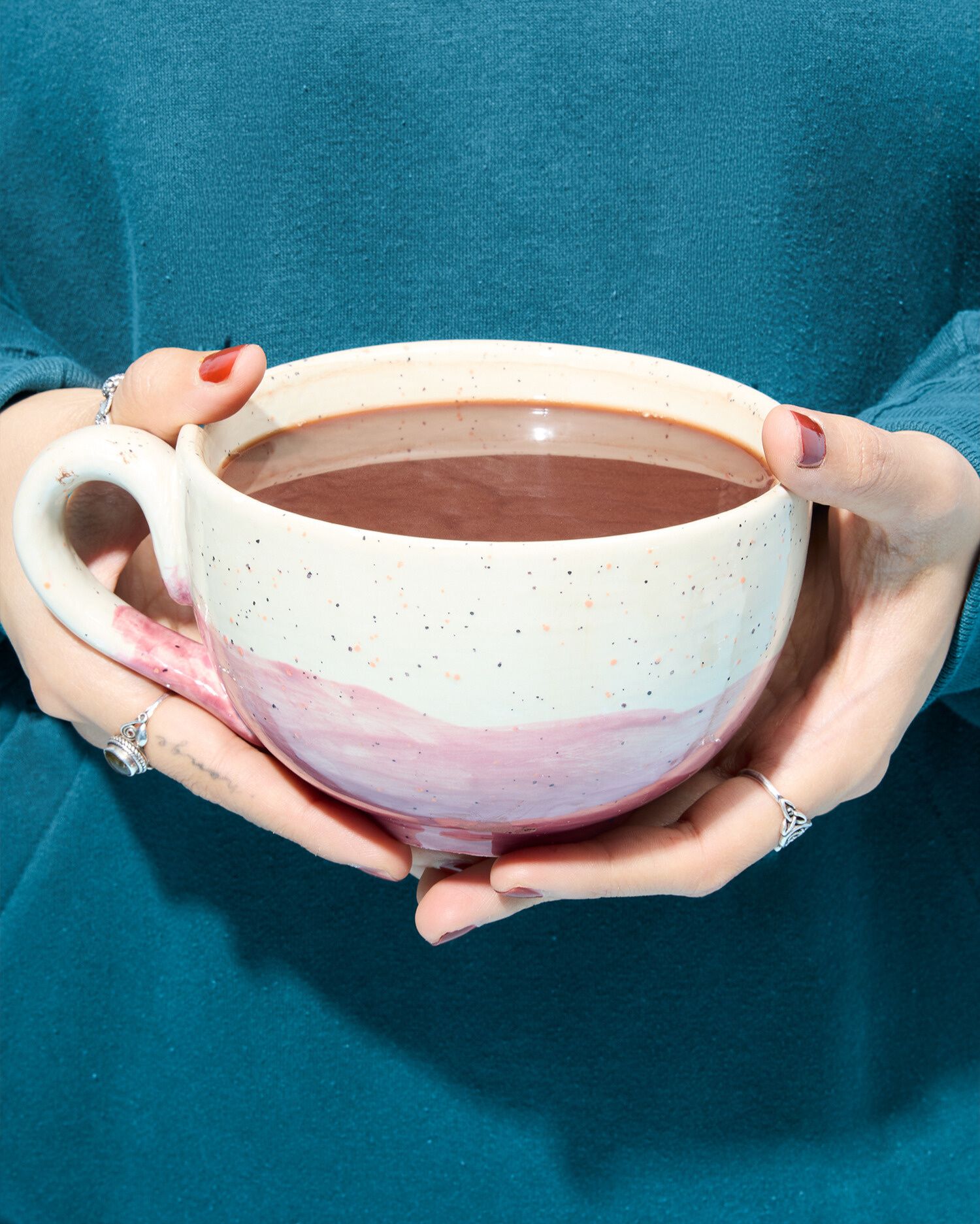 Eine Person in einem petrolfarbenen Pullover hält mit beiden Händen eine große, bauchige Keramiktasse mit heißer, dickflüssiger Trinkschokolade. Die Tasse ist hell, hat dunkle Sprenkel und eine breite, rosafarbene Bemalung im unteren Bereich. Die Person trägt Ringe an den Fingern und hat dunkelrot lackierte Fingernägel.