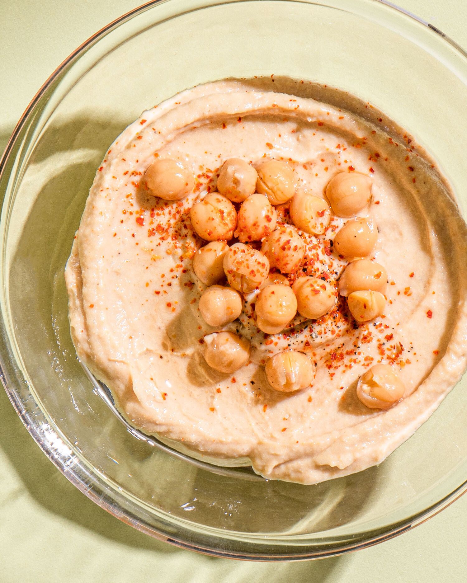 Nahaufnahme von cremigem, hellbraunem Hummus in einer durchsichtigen Glasschüssel, drapiert auf einer hellgrünen Oberfläche. Der Hummus ist in der Mitte mit ganzen, gekochten Kichererbsen und einer Prise Chiliflocken garniert.