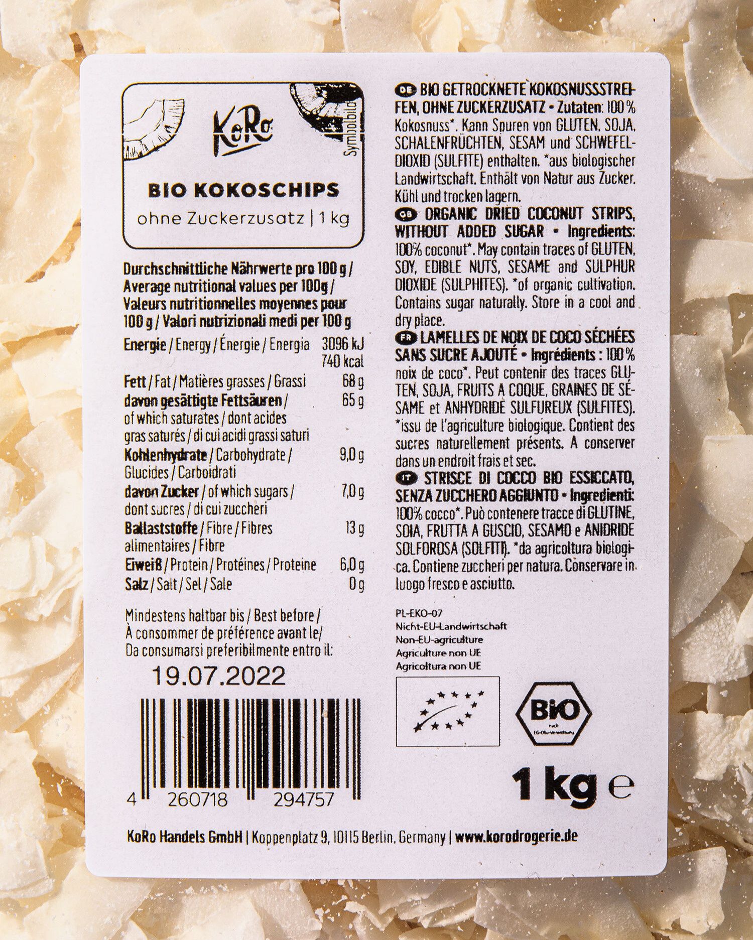 Nahaufnahme einer Produktetikette von KoRo Bio Kokoschips ohne Zuckerzusatz. Die Etikette zeigt die Nährwertangaben, Zutatenlisten in mehreren Sprachen und das Bio-Siegel. Im Hintergrund sind  Kokoschips zu sehen.