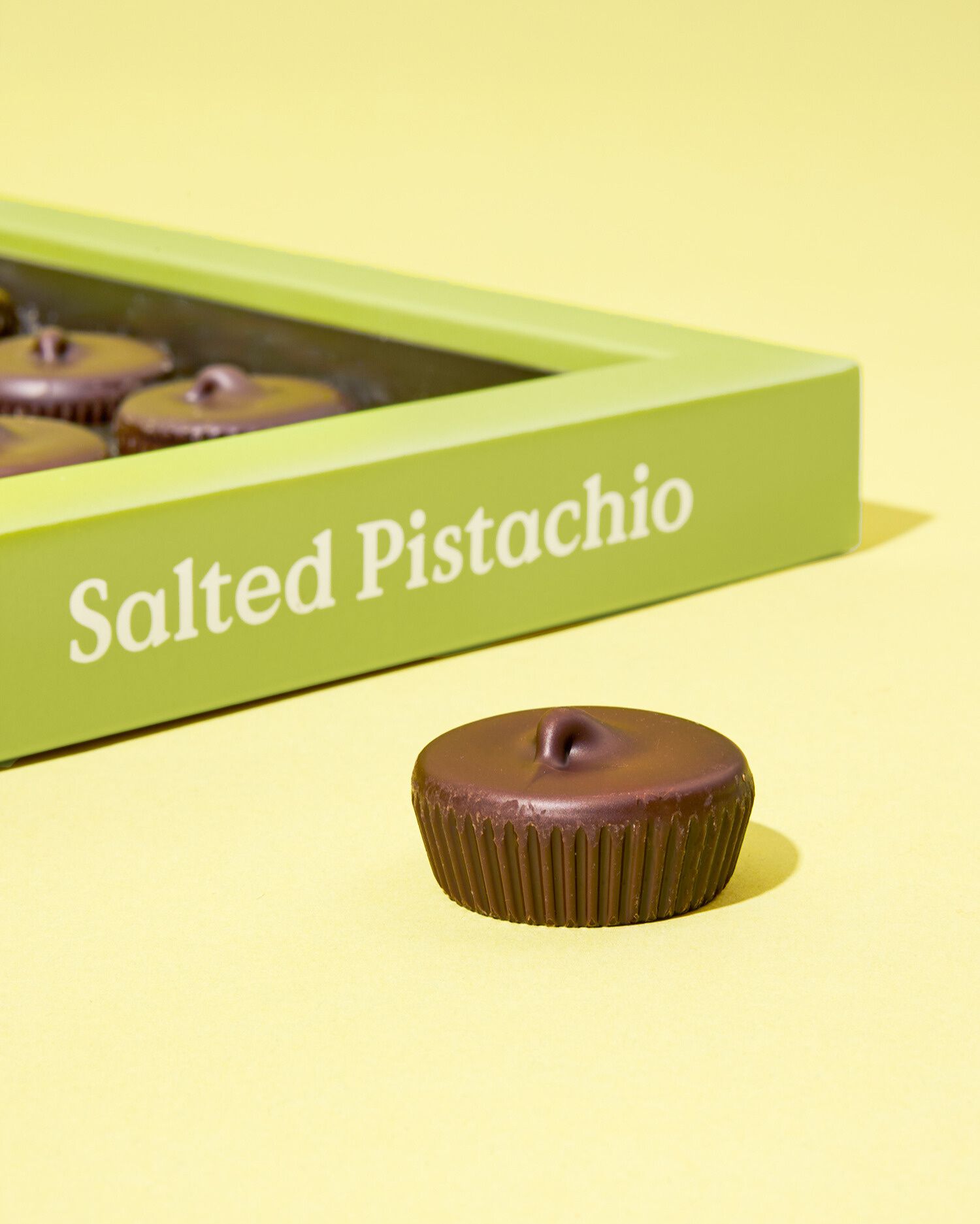 Eine Schokoladenpraline mit geriffeltem Rand liegt vor einer hellgrünen Verpackung mit der Aufschrift ‚Salted Pistachio‘. Der Hintergrund ist in einem passenden hellgelben Ton gehalten.