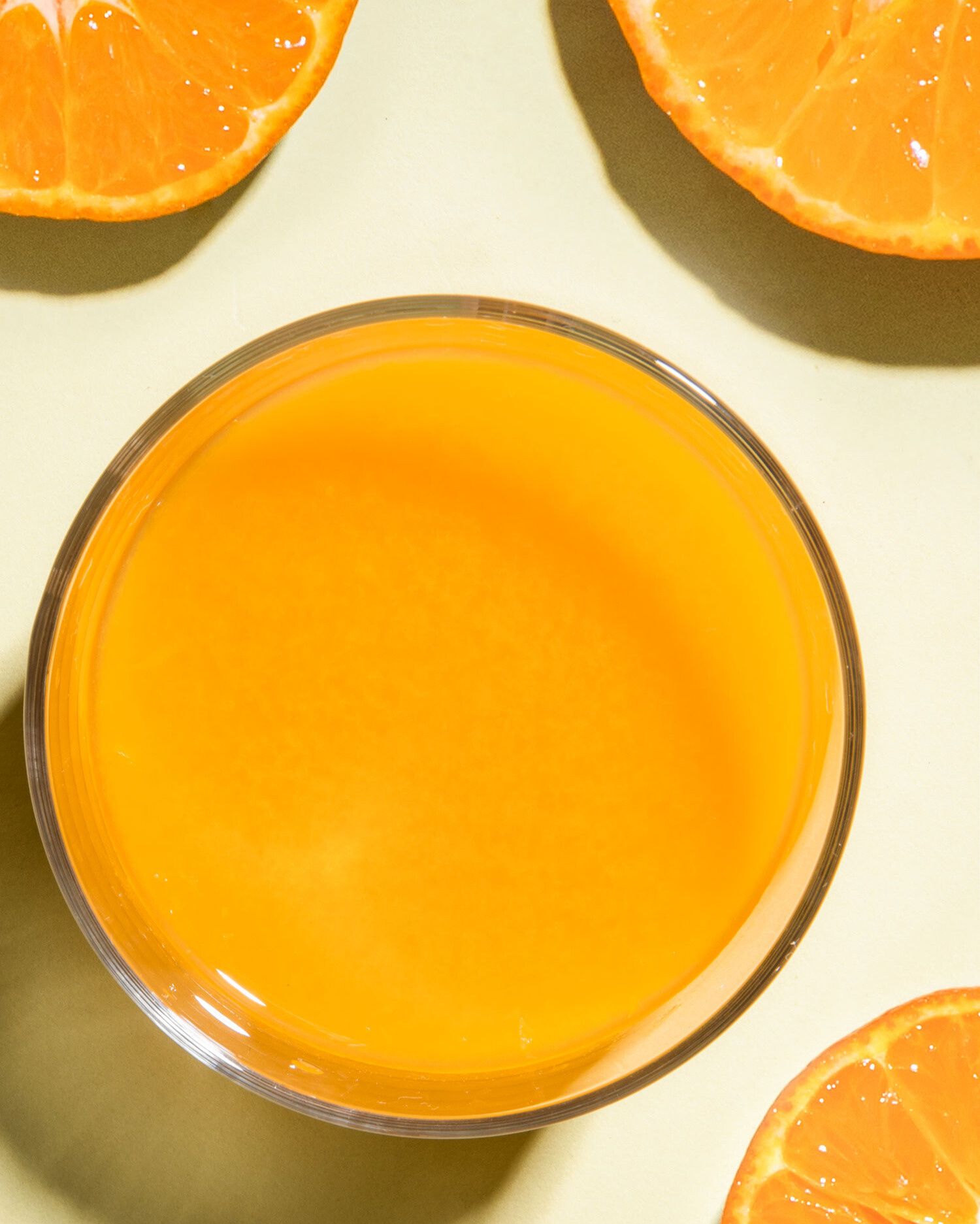 Eine Draufsicht auf ein Glas mit leuchtend orangefarbenem Saft, das von mehreren frischen Zitrusfrüchten auf einem hellgrünen Untergrund umrahmt wird.