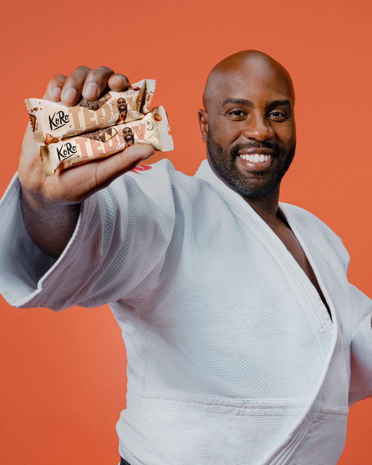Der französische Judoka Teddy Riner lächelt in seinem weißen Judo-Gi und hält zwei KoRo-Proteinriegel seiner eigenen Edition in die Kamera. Der Hintergrund ist in einem kräftigen Orange gehalten.