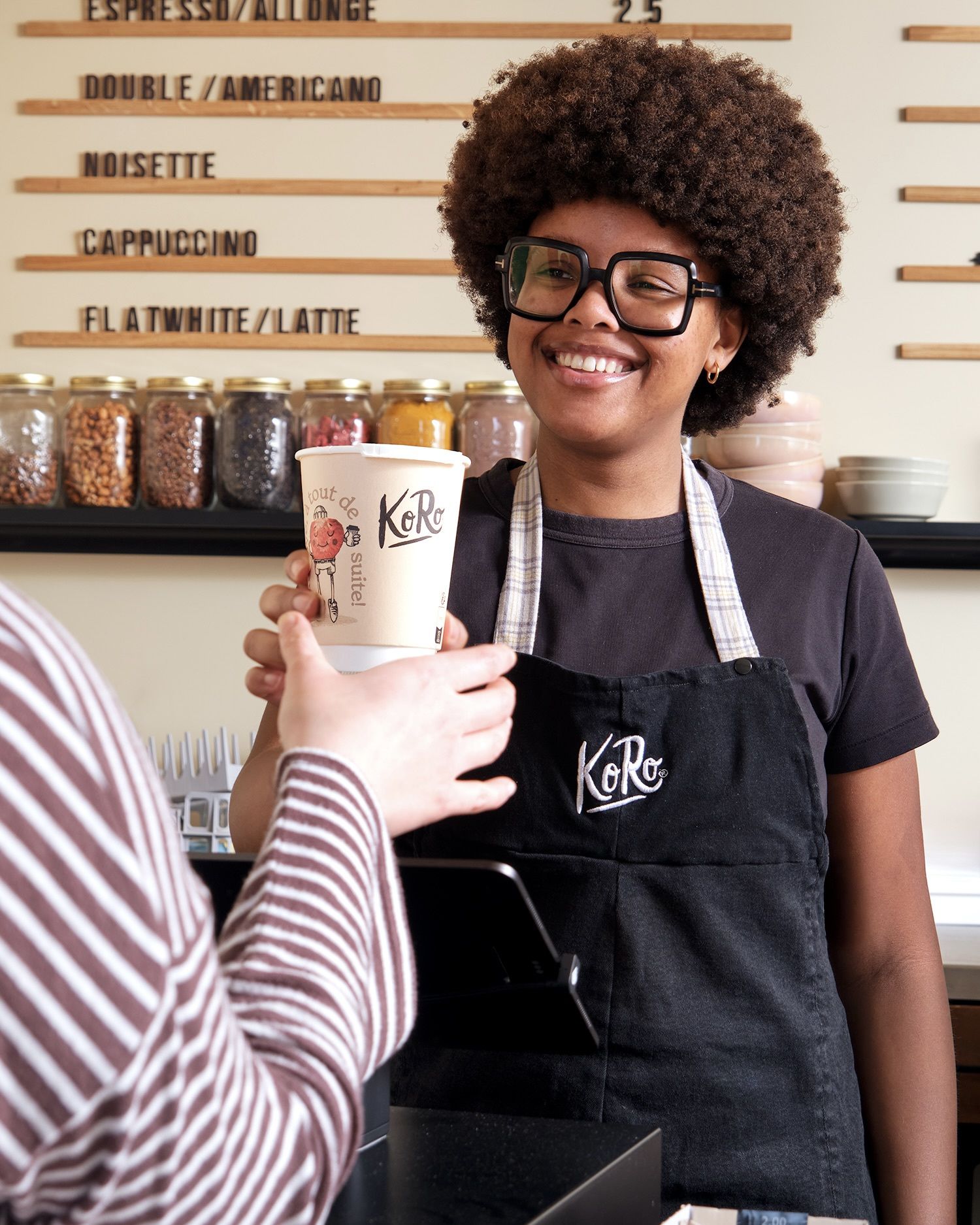 Eine lächelnde Barista mit Brille und Koro-Schürze reicht einem Kunden im KoRo Café einen Kaffeebecher zum Mitnehmen. Im Hintergrund ist die Menütafel mit Kaffeespezialitäten wie Espresso und Cappuccino sowie Gläser mit Zutaten zu sehen.