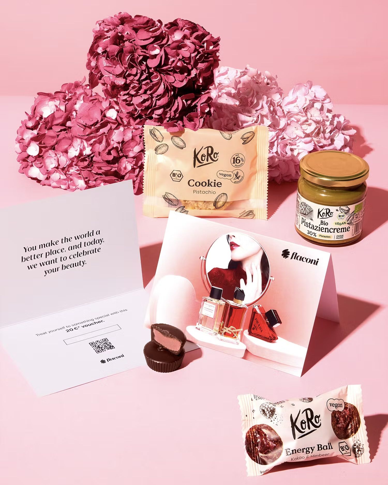 KoRo Muttertags-Geschenkbox mit Pistazien-Cookie, Bio-Pistaziencreme, Nut Butter Cup Cashew Strawberry und einem 20€ Flaconi-Gutschein vor rosa Blumen.