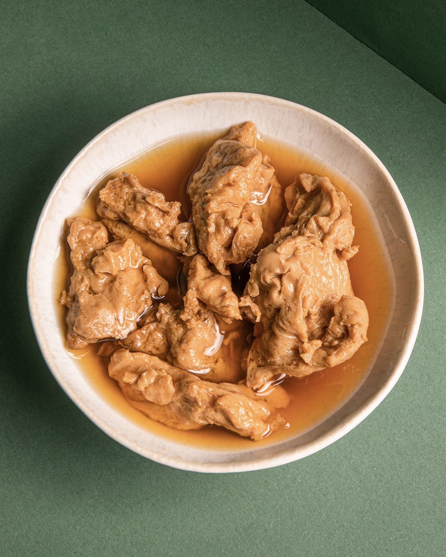 Veganer Fleischersatz: Seitan-Stücke in einer Flüssigkeit in einer Keramikschüssel, fotografiert von oben auf einem grünen Hintergrund.