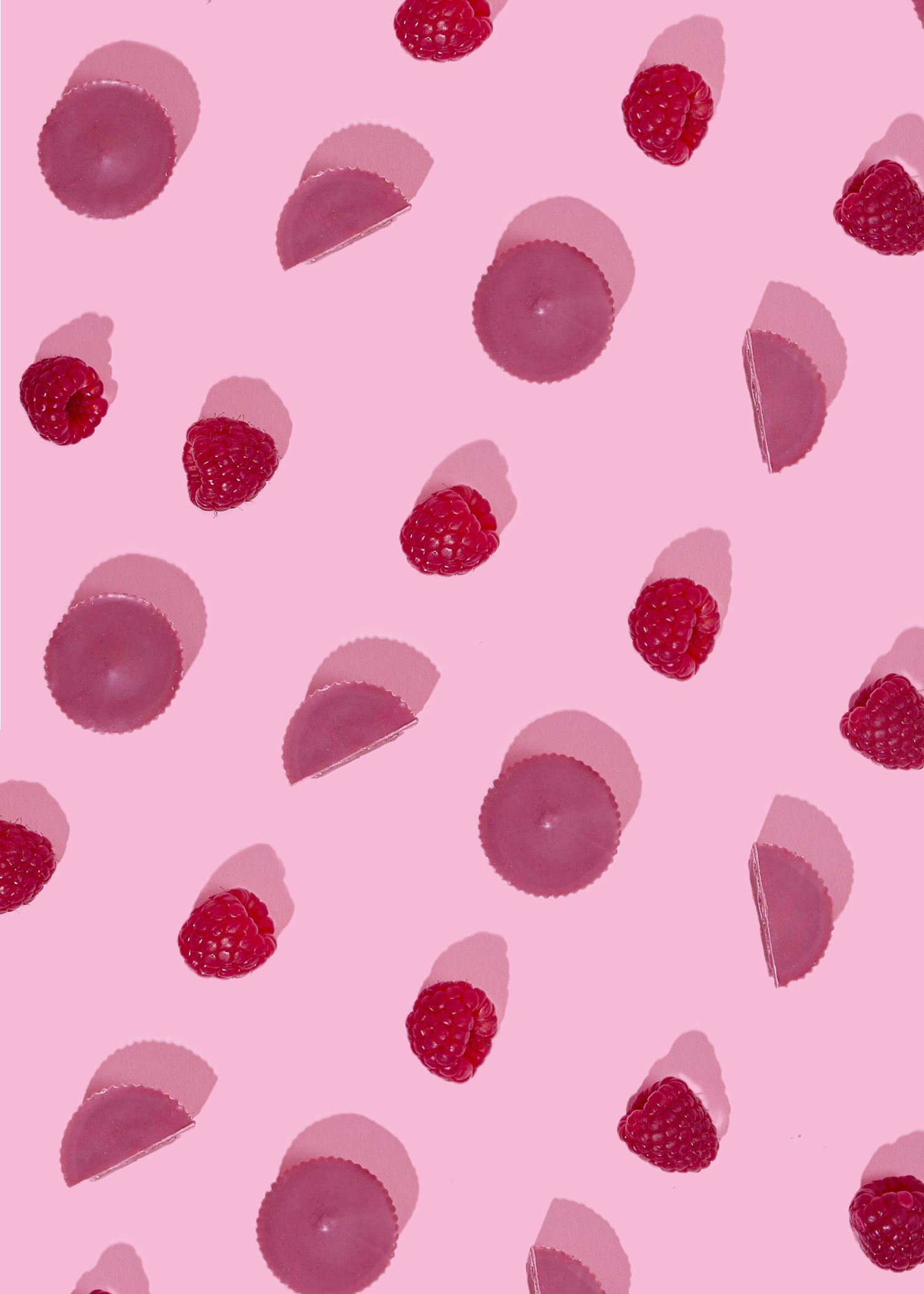 Pattern-Ansicht von oben auf pinkem Hintergrund: Abwechselnd angeordnete Bio Nut Butter Cups Double Raspberry, halbierte Cups und frische rote Himbeeren mit harten Schlagschatten.