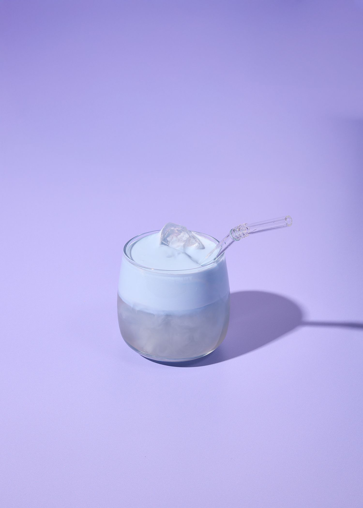 Ein geschichtetes Spirulina-Coconut-Cloud-Getränk in einem klaren Glas mit einer cremigen, pastellblauen Schaumkrone, Kokoswasser und Eiswürfeln, serviert mit einem Glasstrohhalm auf einem minimalistischen, lila Hintergrund.