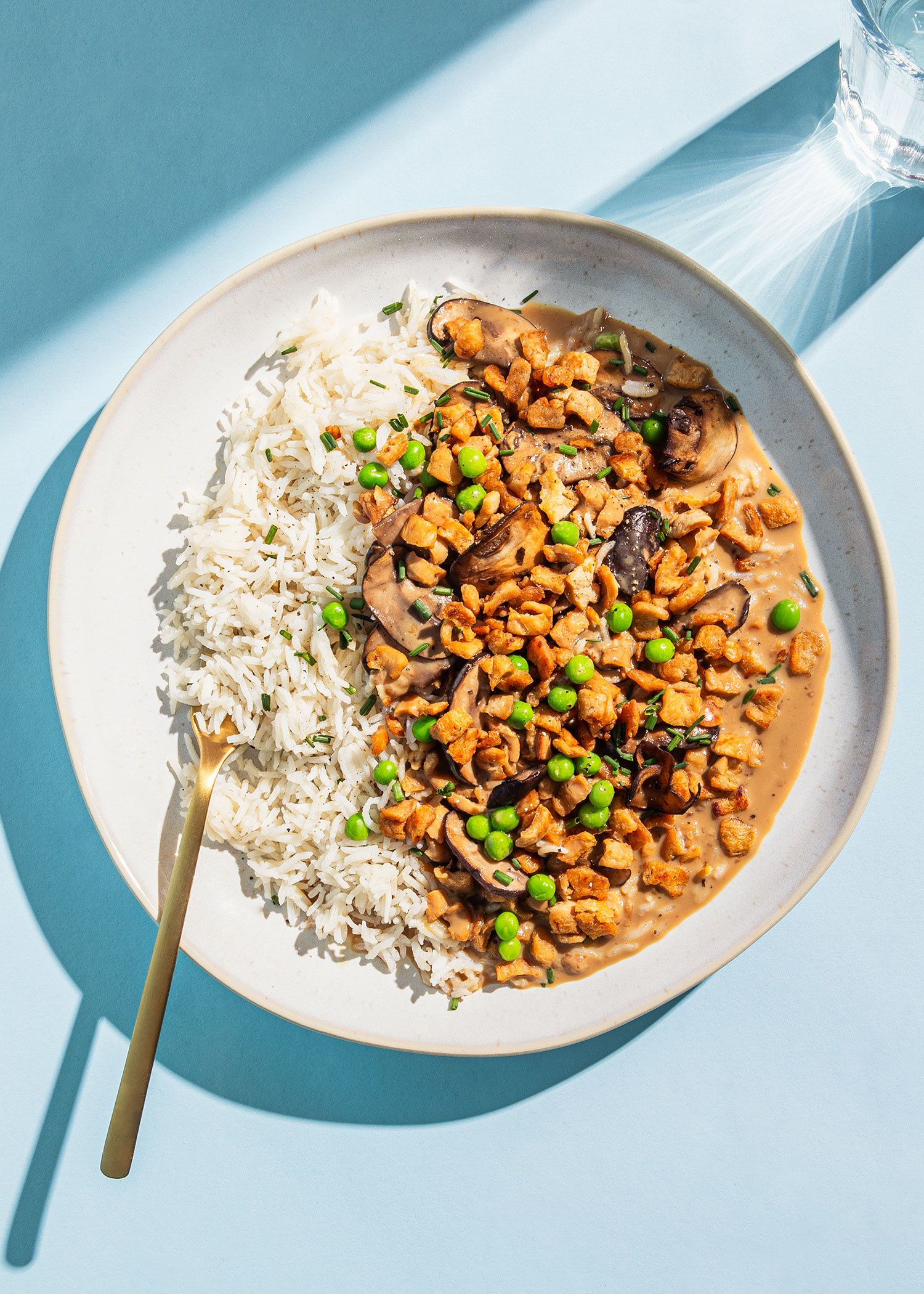 Vogelperspektive auf einen Teller mit weißem Reis und Erbsen-Ackerbohnen-Geschnetzeltem in cremiger Sauce mit Pilzen und Erbsen vor hellblauem Hintergrund.