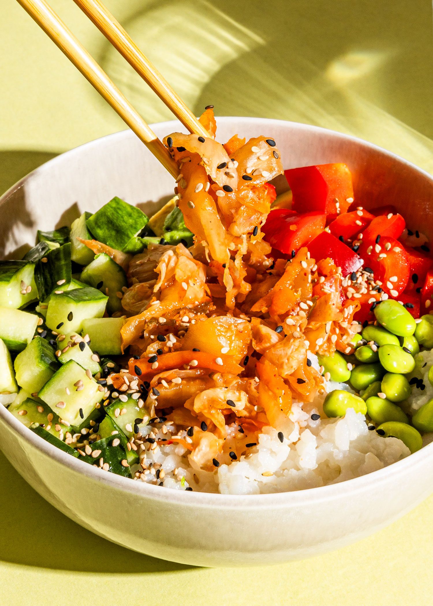 Gemini hat gesagt Eine bunte Bowl mit Reis, Gurken, Paprika und Edamame, aus der mit Holzstäbchen eine Portion Kimchi gehoben wird, angerichtet auf hellgrünem Grund.