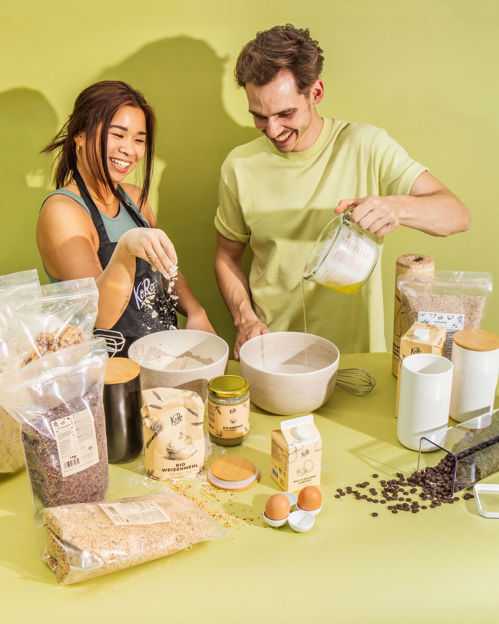  Ein Mann und eine Frau lachen, während sie zusammen backen, vor einem hellgrünen Hintergrund. Die Frau trägt eine schwarze Schürze und streut mit den Fingern etwas in eine Schüssel. Der Mann in einem hellgelben T-Shirt gießt eine Flüssigkeit aus einem Messbecher in eine andere Schüssel. Auf dem Tisch liegen mehrere Kochzutaten, darunter Beutel mit Körnern und Mehl, Eier, Schokoladenstücke, und verschiedene Behälter der Marke KoRo.