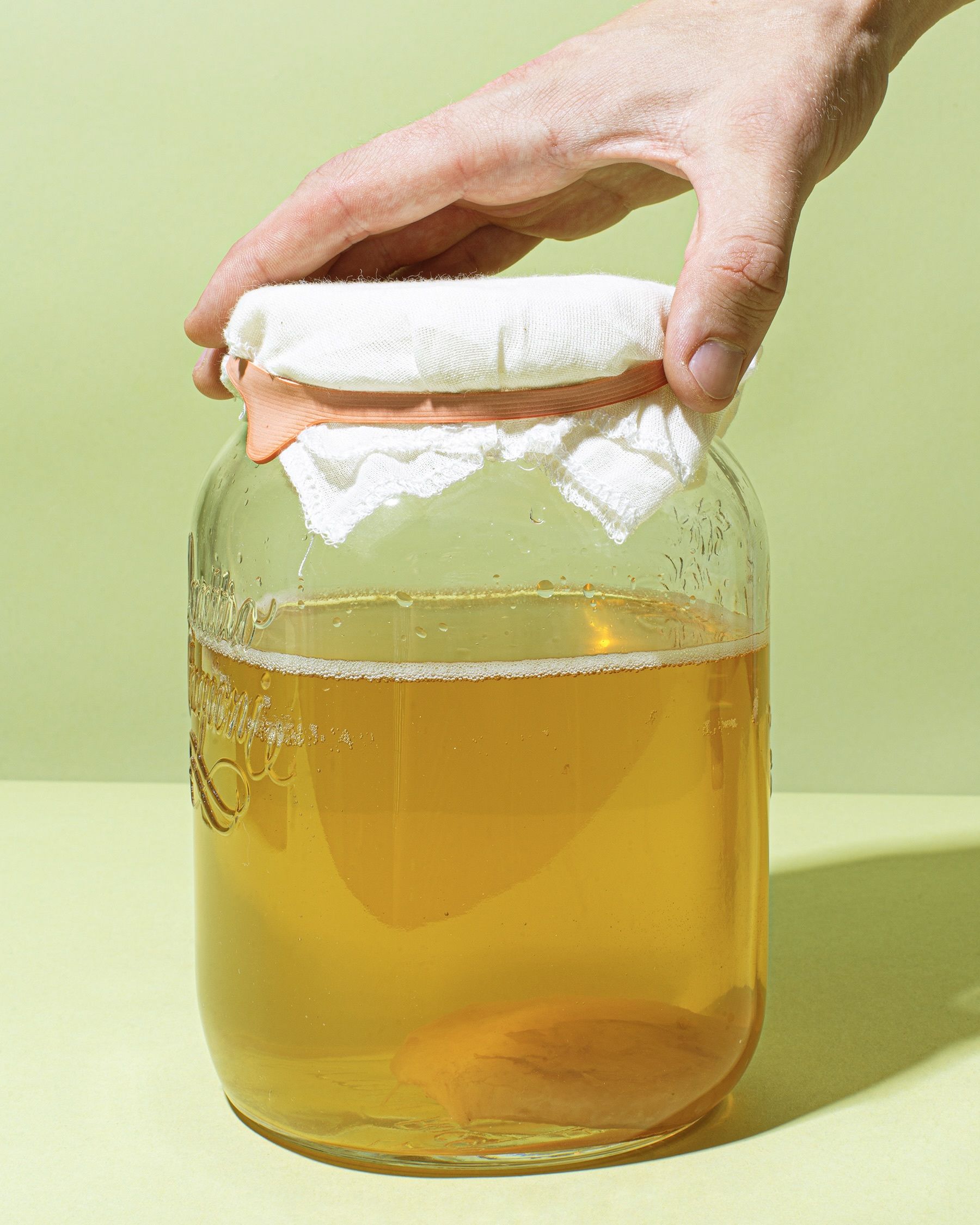 Glas mit Kombucha-Scoby und Starterflüssigkeit als Beispiel für fermentierte Lebensmittel, bedeckt mit einem Tuch und von einer Hand gehalten vor hellgrünem Hintergrund.