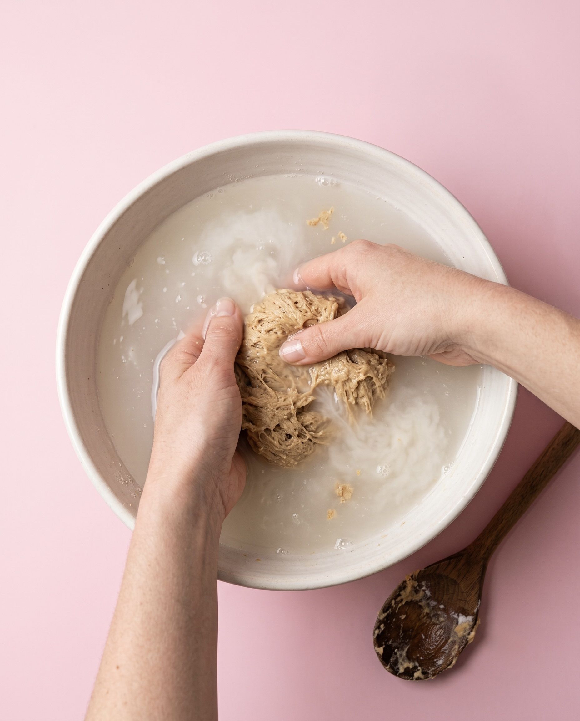 Hände kneten und waschen einen hellbraunen Weizengluten-Teig (Seitan) in einer Keramikschüssel voll trübem Wasser, um die Stärke für die Herstellung von selbstgemachtem veganem Fleischersatz auszuwaschen. Draufsicht vor rosa Hintergrund.