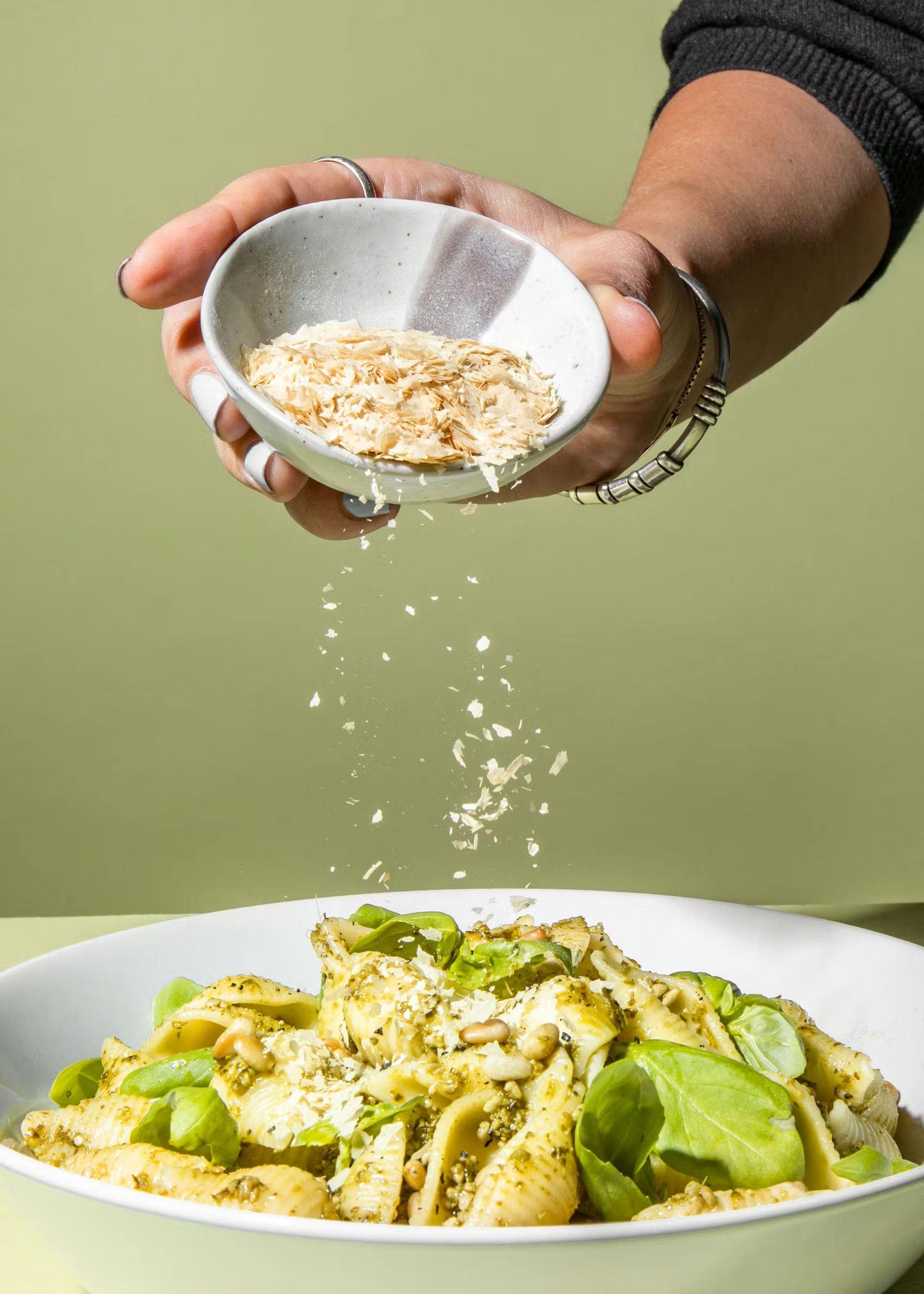 Eine Hand streut goldgelbe Hefeflocken aus einer kleinen Schale über einen Teller Pasta mit grünem Pesto und frischem Basilikum.