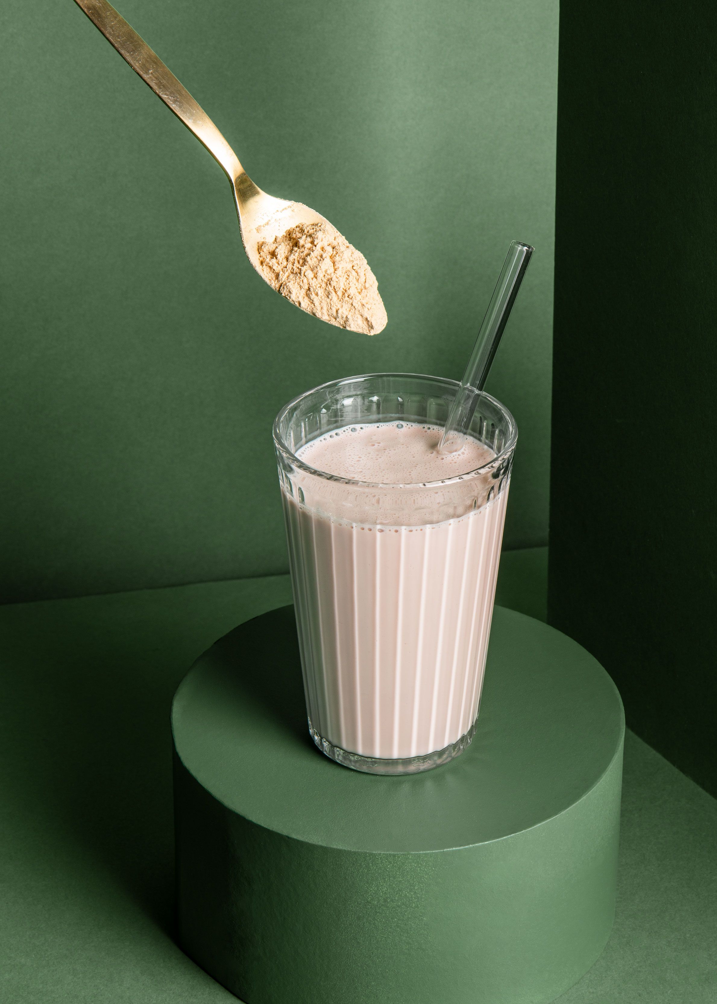 Ein goldener Löffel mit beigem Maca-Pulver schwebt über einem Glas mit einem cremigen, hellbraunen Drink (Maca-Kaffee). Das Glas steht auf einem dunkelgrünen Podest vor einem farblich passenden, grünen Hintergrund.