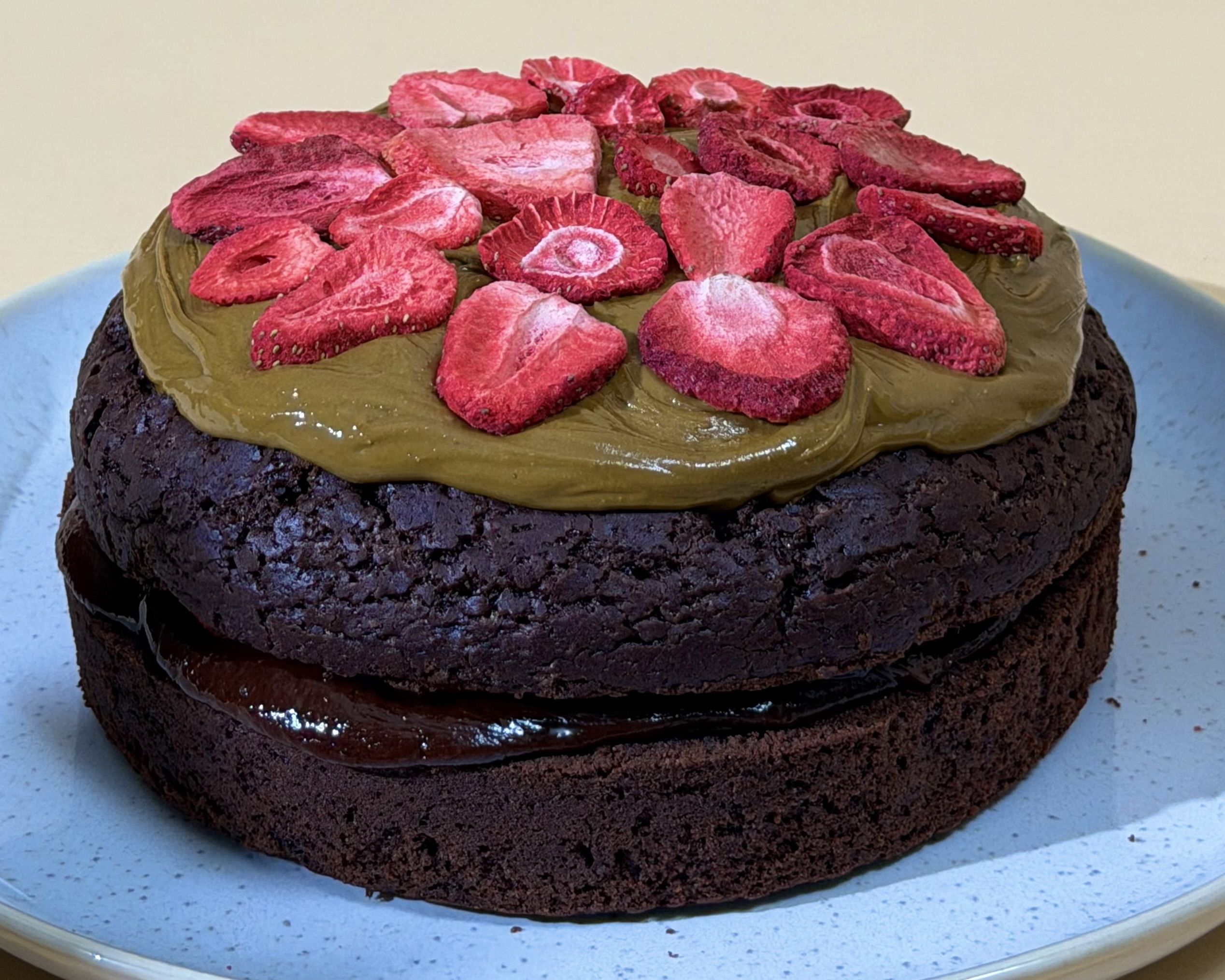 Vegane Schokoladentorte mit Datel Haselnusscreme, oben mit Pistaziencreme bestrichen und mit gefriergetrockneten Erdbeerscheiben dekoriert.