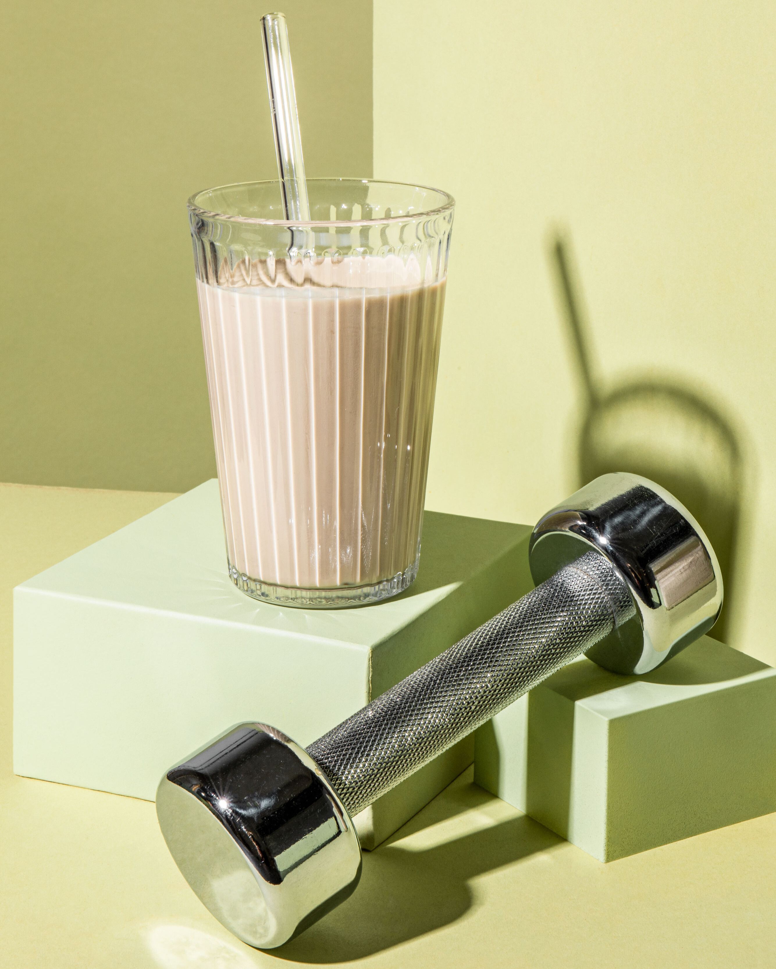 Vor einem grünen Hintergrund steht ein Trinkglas mit einem dickflüssigen Protein-Shake. Daneben liegt eine silberne Hantel.