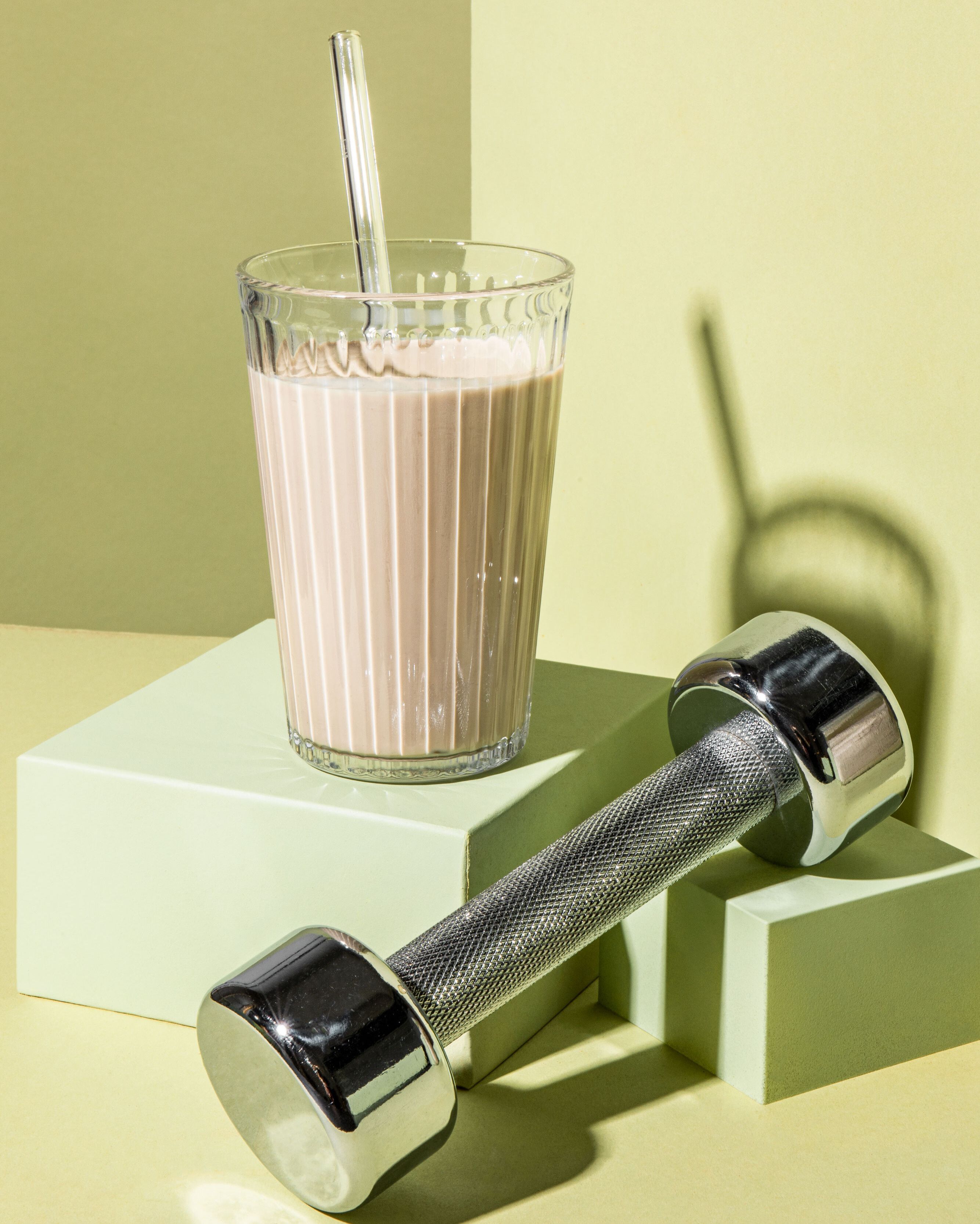 Ein geriffeltes Glas, gefüllt mit einem beigen Protein-Shake, in dem ein durchsichtiger Trinkhalm steckt, steht auf einem hellgrünen, würfelförmigen Podest neben einer glänzenden, silbernen Hantel. Das Arrangement steht vor einem hellgelb-grünen Hintergrund.