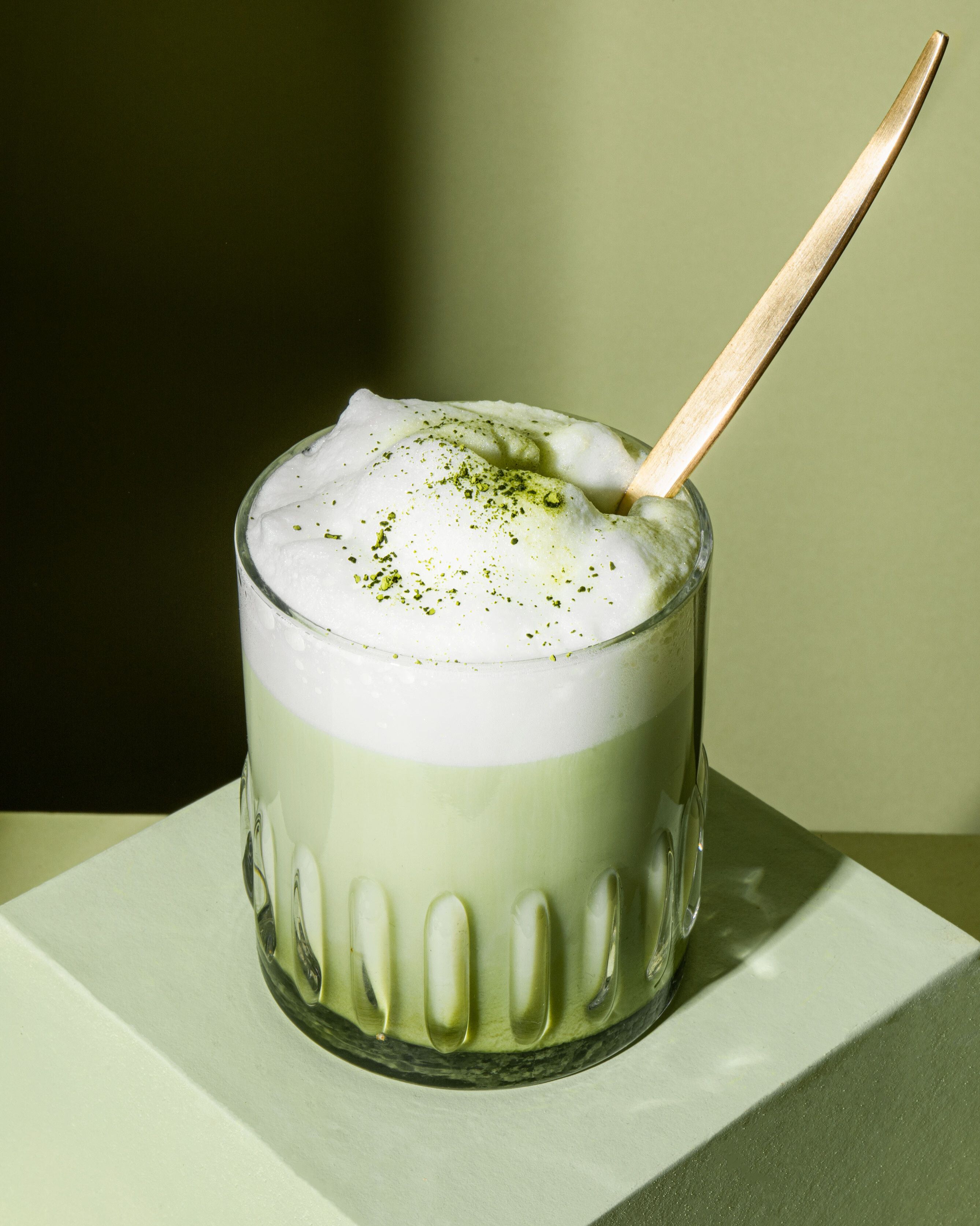 Ein durchsichtiges, geriffeltes Glas mit einem grünen Matcha-Latte, der eine Schicht aus dickem, weißem Schaum und ein paar Matcha-Pulver-Sprenkeln darauf hat. 