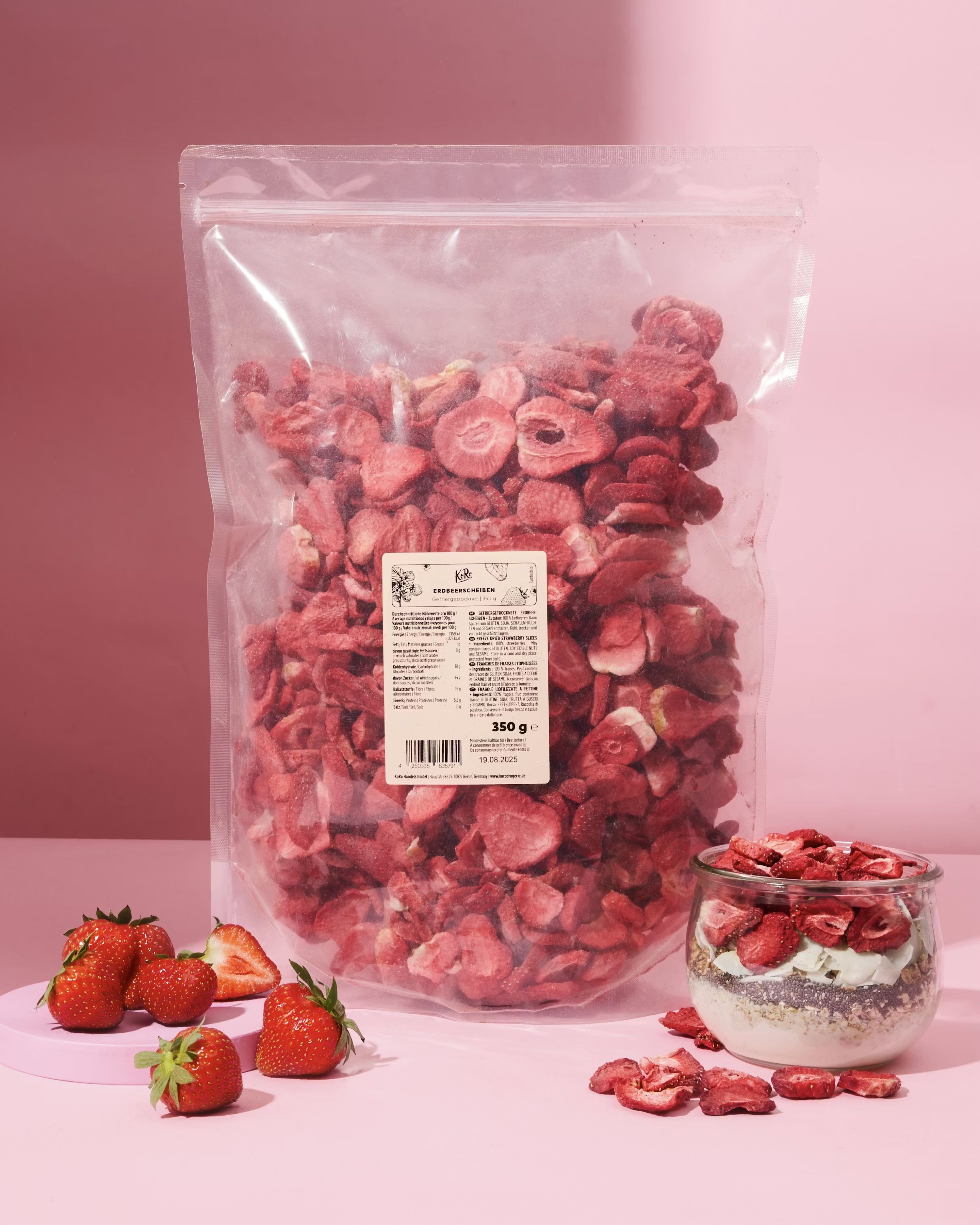 Große 350g-Packung gefriergetrocknete Erdbeerscheiben von KoRo, zusammen mit frischen Erdbeeren und einem dekorierten Müsli-Glas vor einem rosa Hintergrund.