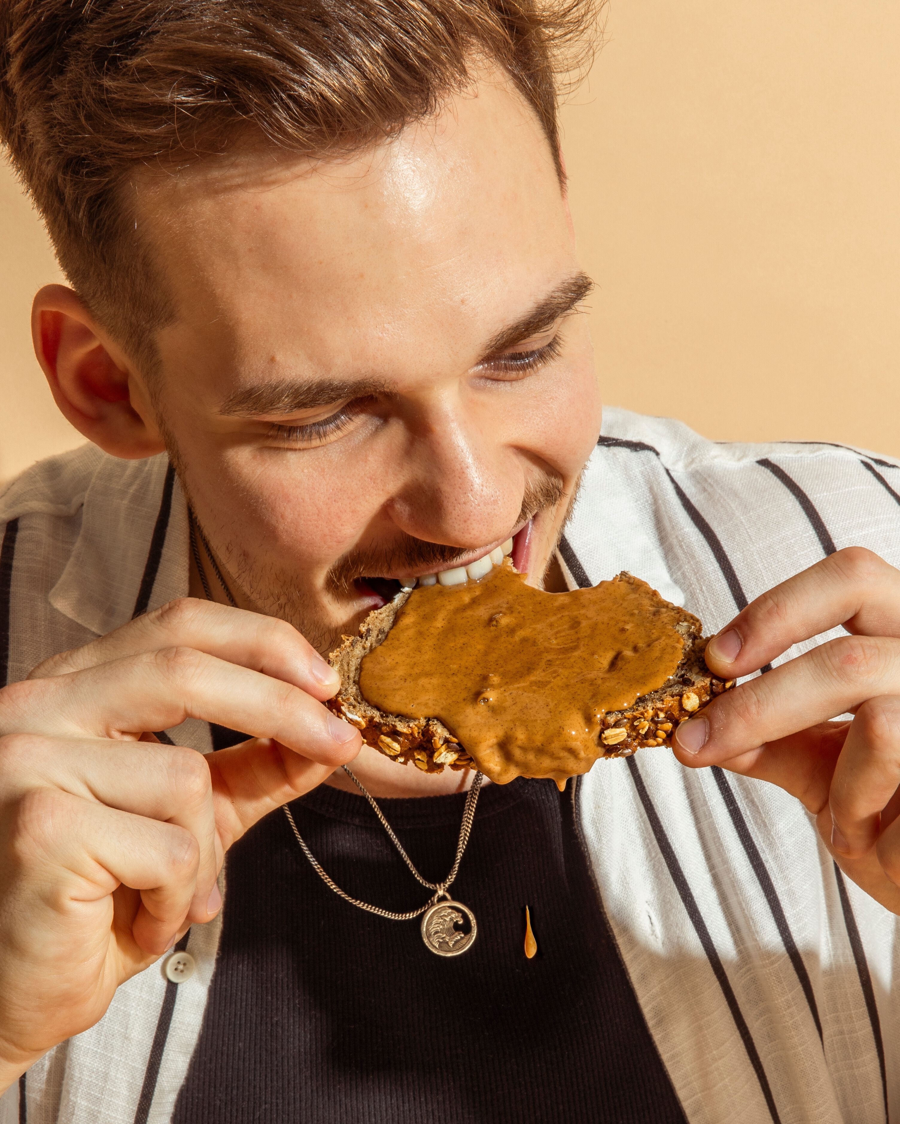 Ein junger Mann mit braunem Haar und gestreiftem Hemd beißt herzhaft in eine Scheibe Vollkornbrot, die großzügig mit cremigem Mandelmus bestrichen ist. Er lächelt dabei und hält das Brot mit beiden Händen vor einem hellen, warmen Hintergrund.
