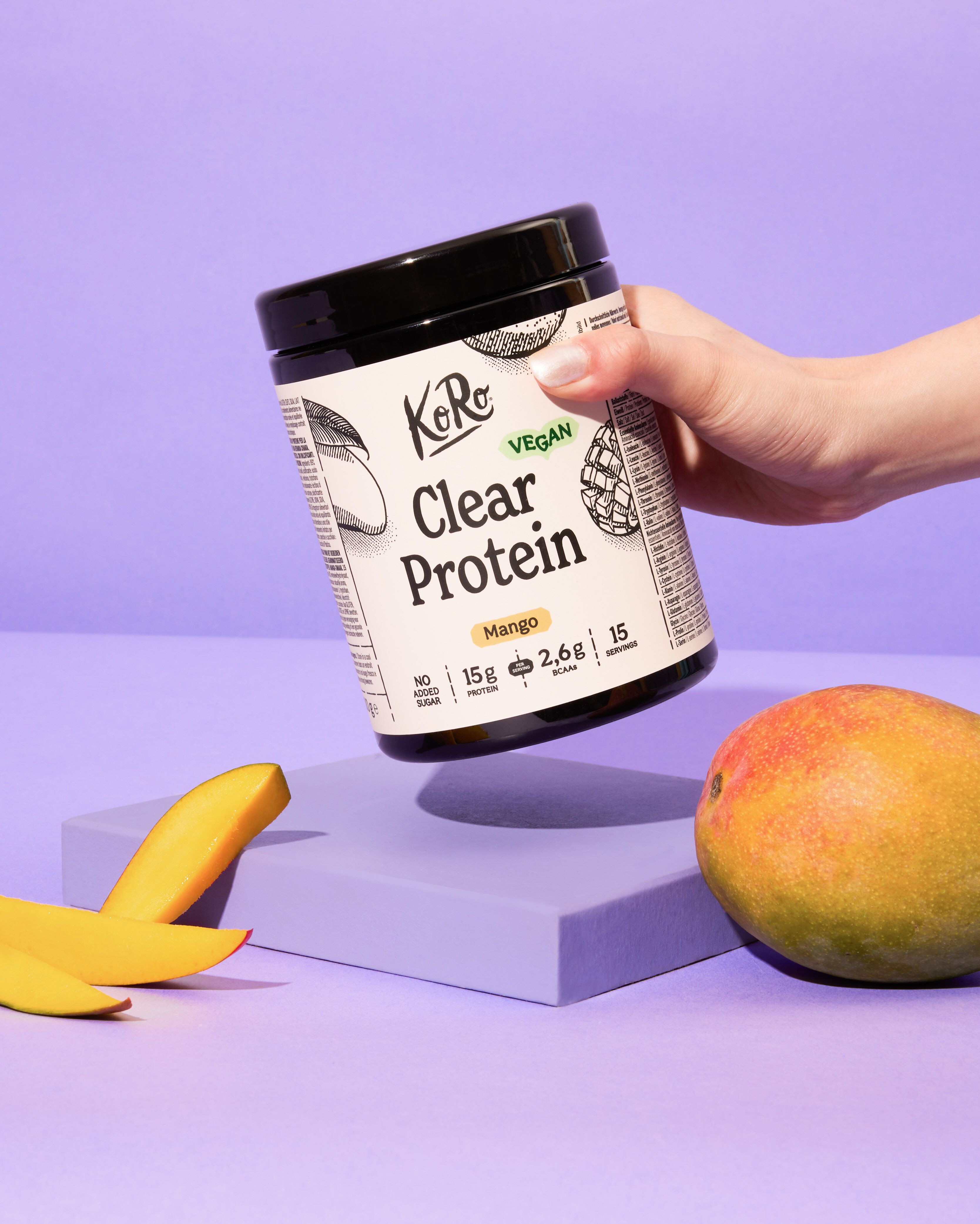 Hand hält ein schwarzes Dose mit weißem Etikett von KoRo Clear Protein in der Geschmacksrichtung Mango vor lilafarbenem Hintergrund. Neben der Dose liegen eine ganze Mango und zwei Mangoscheiben auf einer lilafarbenen Oberfläche.