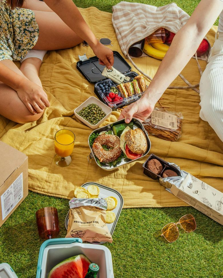 Zwei Personen sitzen bei sonnigem Wetter auf einer gelben Picknickdecke im Gras und teilen sich eine vielfältige Auswahl an Speisen, darunter belegte Bagels, Sandwiches, frische Beeren, Edamame und Snacks in verschiedenen Boxen und Verpackungen.