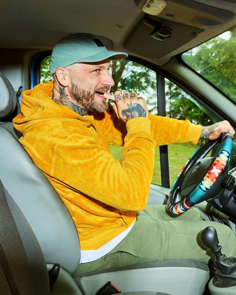 Ein tätowierter Mann mit Bart und Basecap sitzt lächelnd am Steuer eines Autos, hält das bunt gemusterte Lenkrad und isst während der Fahrt einen Snack.