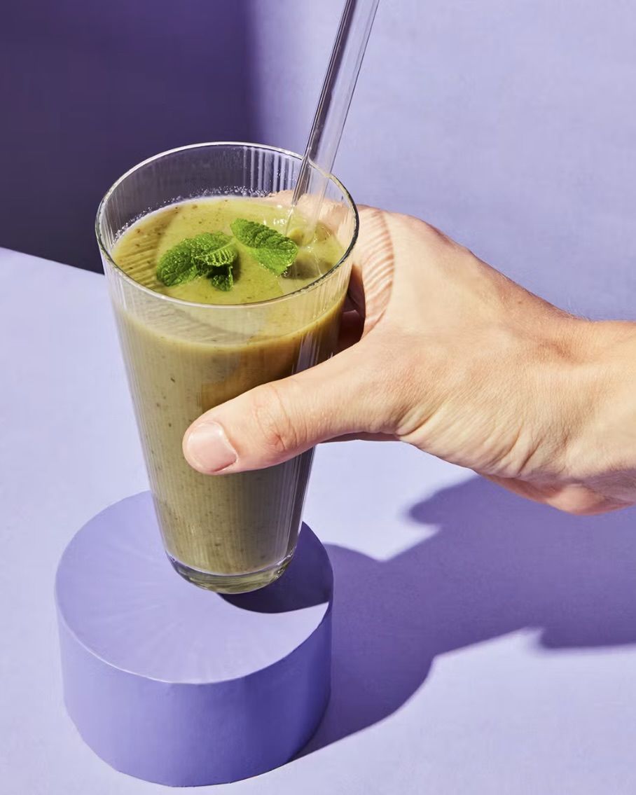 Eine Hand hält ein Glas mit einem vitalisierenden Superfood-Smoothie aus grünem Pulver und Minzblättern vor einem minimalistischen, lila Hintergrund.
