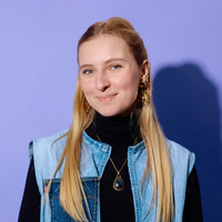 Flavie lächelt in die Kamera, sie trägt ein schwarzes Oberteil und eine Jeansweste sowie große Ohrringe und eine Kette