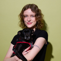 Lisa steht lächelnd vor der Kamera und hat einen schwarzen Hund auf dem Arm, der ebenfalls in die Kamera schaut