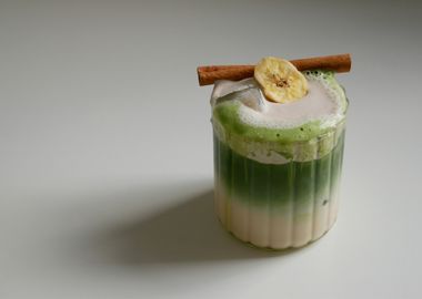 Ein Glas auf einem weißen Tisch, gefüllt mit einem Getränk. Unten ist eine milchige Flüssigkeit, darüber eine Schicht aus leuchtend grünem Matcha-Tee. Das Getränk ist mit Eiswürfeln versehen und hat eine Schaumkrone, die bis über den Rand läuft. Auf dem Schaum liegen eine Zimtstange und eine getrocknete Bananenscheibe.