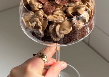 Ein Close-up-Foto zeigt einen dunklen Schokoladen-Brownie-Kuchen, der in einem eleganten Martiniglas serviert wird. Der Kuchenboden ist mit einer großzügigen Schicht einer cremigen, hellbraunen Masse bedeckt, in die knusprige, kakaofarbene Cerealien gemischt sind. Das Glas wird von unten von einer Hand mit einem goldenen Ring gehalten, die einen schwarzen Ärmel trägt.