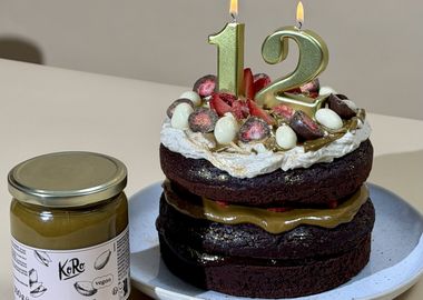 Veganer Schokoladen-Geburtstagskuchen mit goldenen Zahlenkerzen „12“, gefüllt mit Pistazienmus und frischen Erdbeeren, dekoriert mit Sahne, halbierten Schoko-Erdbeeren und weißen Schoko-Eiern auf einem hellen Teller; daneben ein Glas KoRo Pistazienmus – festlich dekorierte Geburtstagstorte zum 12. Geburtstag.