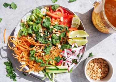 Thai Nudelsalat mit cremiger Erdnusssauce, frischem Gemüse, Koriander, Limettenspalten und gerösteten Erdnüssen in einer Schüssel