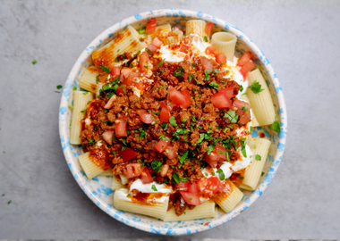 Ein Teller mit türkischer Pasta (Manti-Style): Große Rigatoni-Nudeln getoppt mit cremigem Knoblauchjoghurt, würzig angebratenem veganen Hackfleisch, frischen Tomatenwürfeln und gehackter Petersilie in einer blau-weiß gesprenkelten Schüssel.