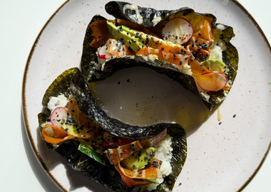 Zwei knusprige Sushi Tacos aus Nori-Blättern, gefüllt mit Sushi-Reis, Avocado, Radieschen, veganer Karotten-Lachs und schwarzer Sesam, angerichtet auf einem hellen Teller im Sonnenlicht.