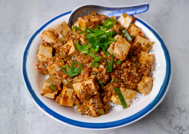 Eine Schüssel serviertes veganes Mapo Tofu: Asiatisches Gericht mit Tofu, Sojagranulat in würziger Sauce und Frühlingszwiebel-Ringen auf einem hellen Hintergrund.