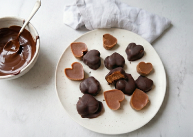 Vegane Nougat Herzen mit Haselnussmus auf einem hellen Teller, überzogen mit Schokoladenglasur aus Xylit-Schokodrops, daneben eine Schüssel mit geschmolzener Schokolade und Löffel auf hellem Marmortisch.